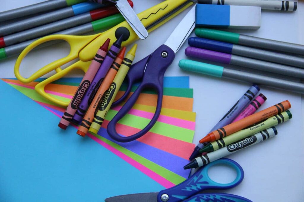Scissors, crayons, erasers, markers on top of construction paper on a desk.
